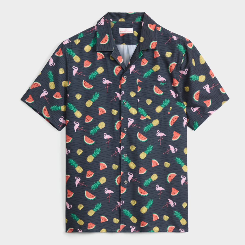 The TROPICAL TANGO SHIRT by PUBLIC BEACH SWIM is a short-sleeve, button-up shirt with a dark background and vibrant pineapples, watermelon slices, and pink flamingos—ideal for vacations or stylish beachwear.