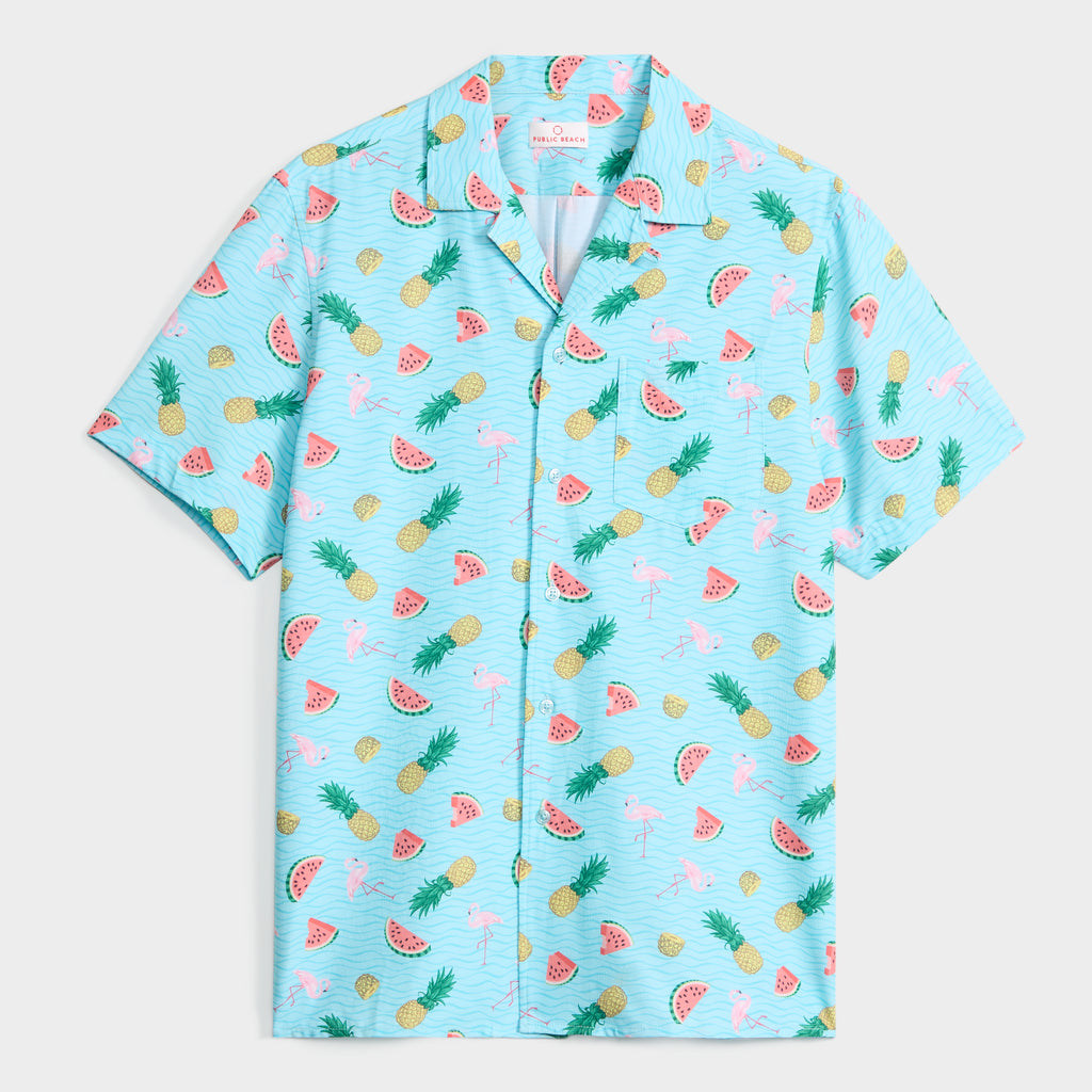 The TROPICAL TANGO SHIRT by PUBLIC BEACH SWIM is a short-sleeve, button-up with a bright blue background and fun pineapple, watermelon, and flamingo prints—ideal for sunny days or beach outings with its relaxed, playful style.