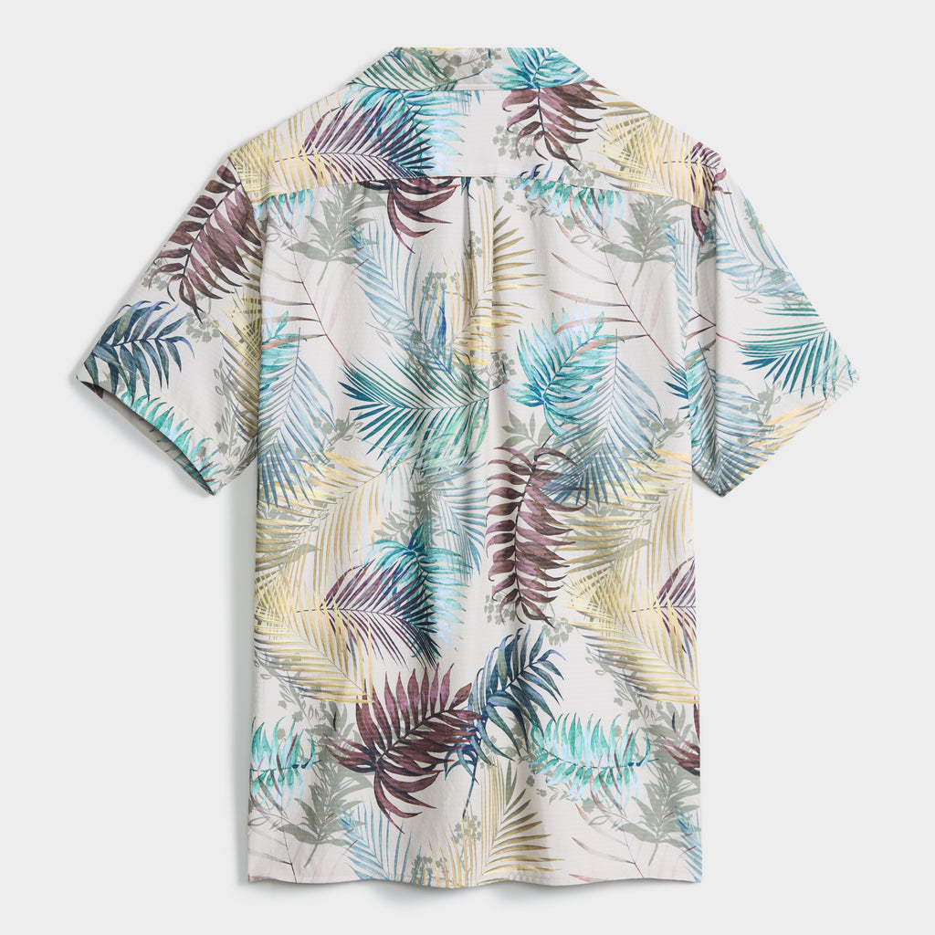 The BOTANIC BURST SHIRT by PUBLIC BEACH SWIM is a men's button-up with a tropical palm and leaf print in green, blue, yellow, and burgundy on a light background—ideal for summer style.