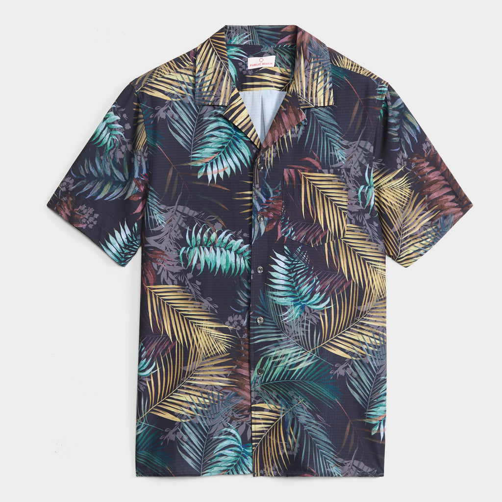 The BOTANIC BURST SHIRT by PUBLIC BEACH SWIM is a short-sleeve men’s button-up with a vibrant green, yellow, and brown palm print on dark fabric—ideal for a fresh summer look.