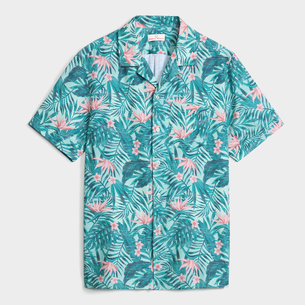 The TROPICAL PULSE SHIRT by PUBLIC BEACH SWIM is a short-sleeve, button-up Cuban-collar shirt with green leaves and pink flowers printed on light seersucker fabric.