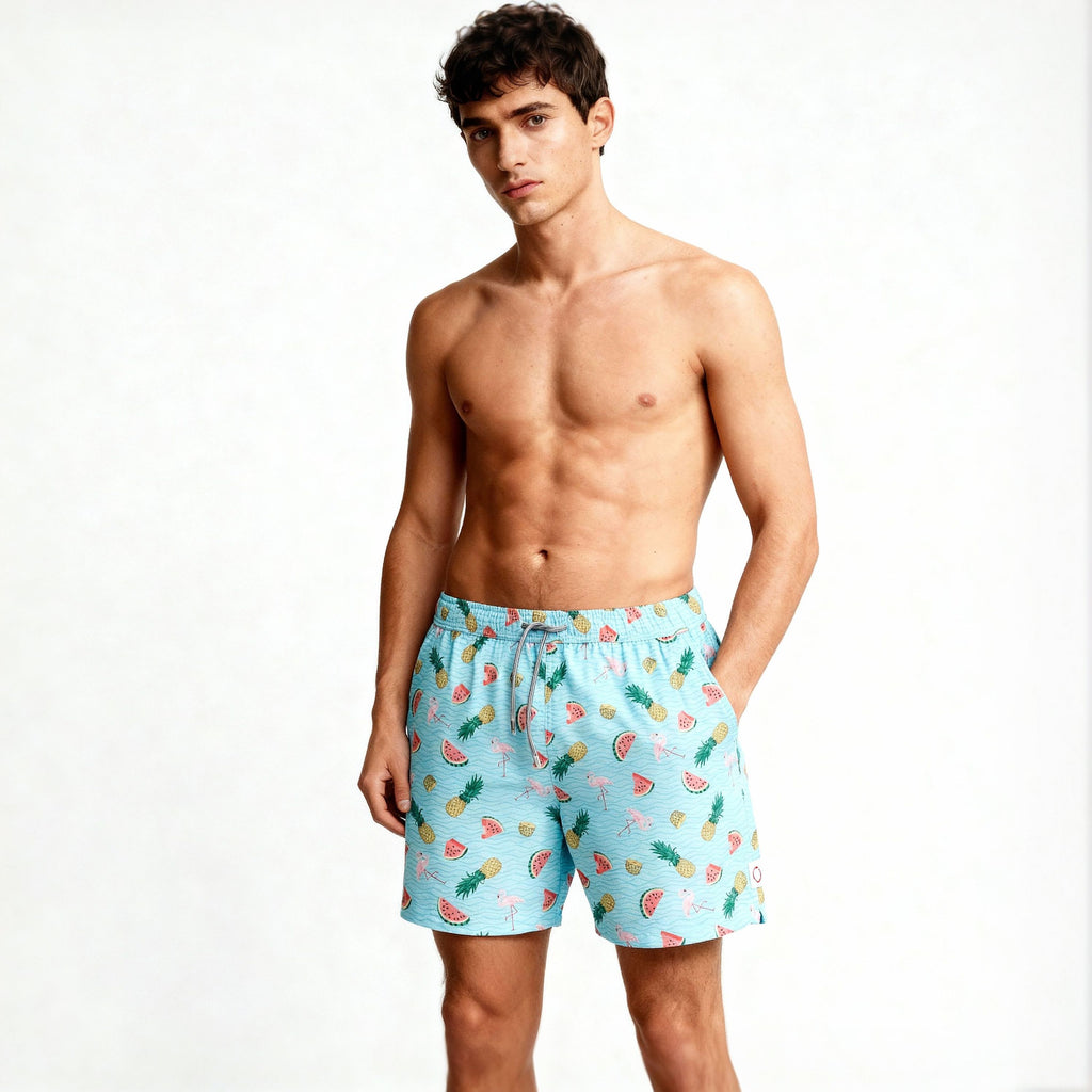 A young man stands shirtless against a plain white background, modeling PUBLIC BEACH SWIM’s TROPICAL TANGO light blue swim shorts, which feature colorful fruit patterns like watermelon, pineapple, and orange slices.