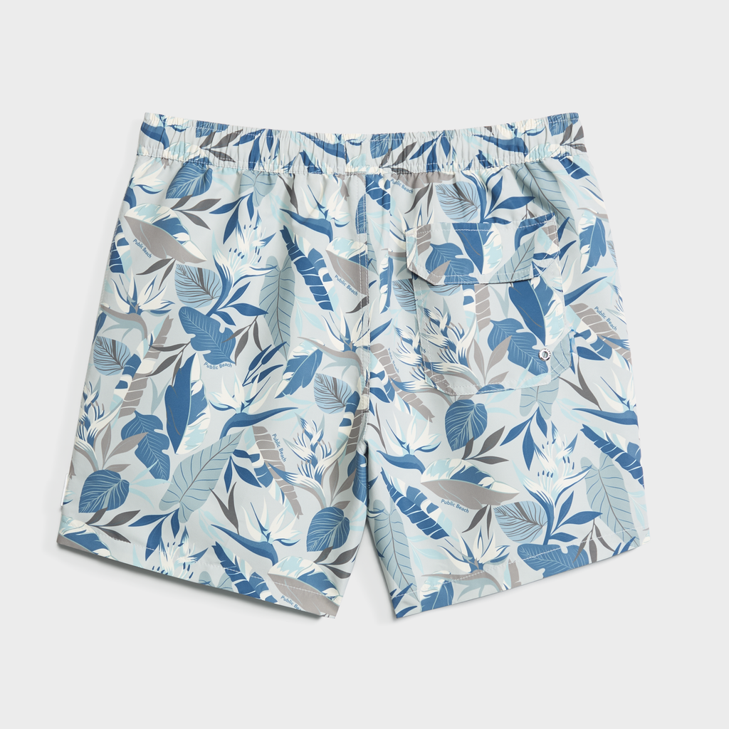 The BLOOMING TROPIC swim trunks by PUBLIC BEACH SWIM feature a blue and white floral print, elastic waistband, and back pocket. Quick-dry and ideal for tropical pool or beach days, they're displayed flat on a white background.