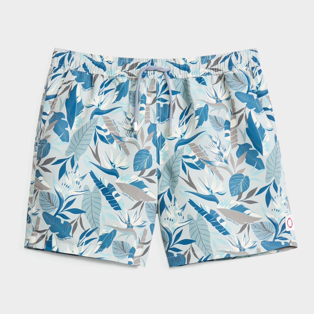 The BLOOMING TROPIC swim trunks by PUBLIC BEACH SWIM feature a light blue, white, and gray leaf print, elastic waistband with drawstring, and a small logo on the right hem. Quick-dry fabric makes them perfect for sunny days.