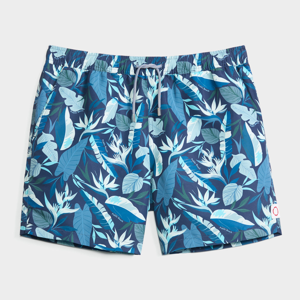 The BLOOMING TROPIC swim trunk by PUBLIC BEACH SWIM features an elastic waistband, drawstring, quick-dry fabric, and a vibrant blue-and-white floral print with leaf and bird of paradise motifs.