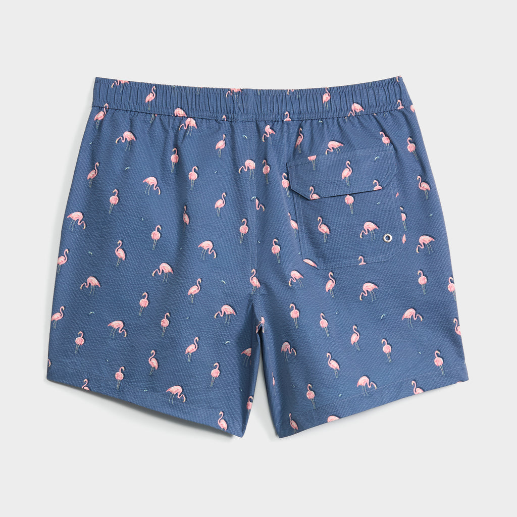 The FLAMINGLOW swim trunk by PUBLIC BEACH SWIM features blue seersucker fabric with small pink flamingos, an elastic waistband, and a back pocket.