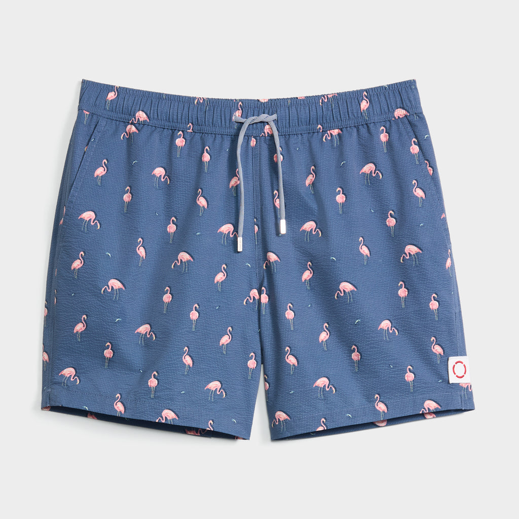 The FLAMINGLOW swim trunk by PUBLIC BEACH SWIM features a blue quick-dry fabric with pink flamingo print, elastic waistband, grey drawstring, and a white patch with red circle on the front left leg.