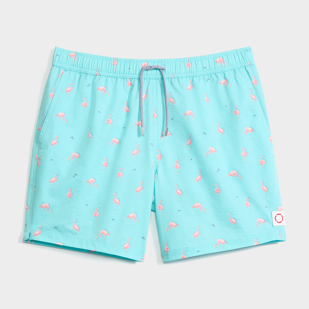The FLAMINGLOW swim trunk by PUBLIC BEACH SWIM features an aqua blue color with a pink flamingo print, elastic waistband with drawstring, and a white patch with red circle. Quick-dry—ideal for sunny days.