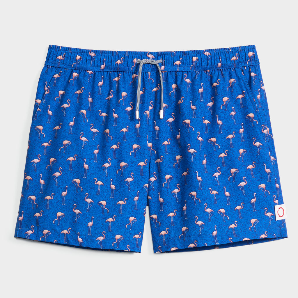 The FLAMINGO VIBES swim trunk by PUBLIC BEACH SWIM features a bright blue print with pink flamingos, white drawstring, quick-dry fabric, and a small white patch with a red circle near the left leg hem.