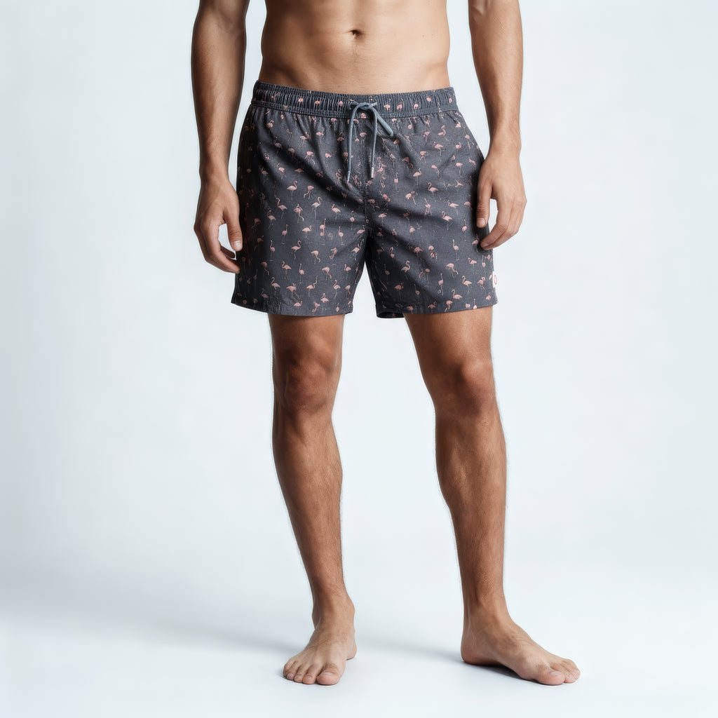 A man stands barefoot against a plain background, wearing PUBLIC BEACH SWIM's FLAMINGO VIBES swim trunks in gray with a pink flamingo pattern. His upper body is bare and hands at his sides—quick-dry swimwear ideal for sunny beach days.