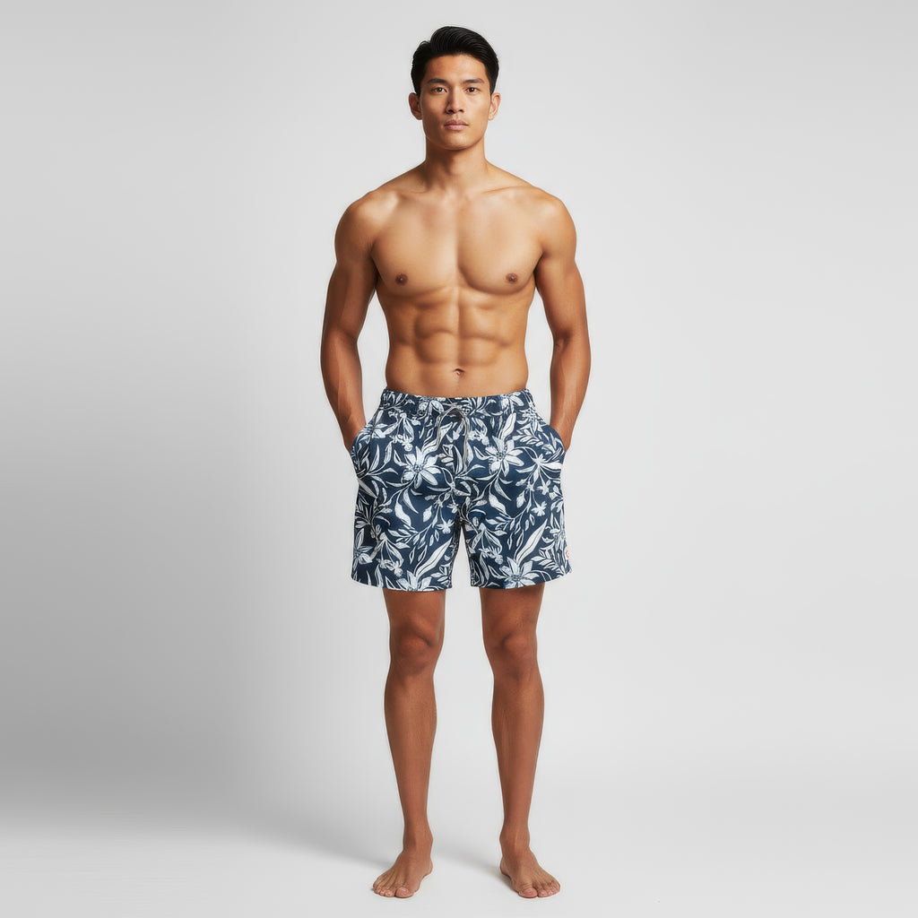 A shirtless man faces forward with hands in the pockets of PUBLIC BEACH SWIM's FLORAL IVY men’s swim shorts, featuring a navy and white floral ivy print, against a plain light gray background.