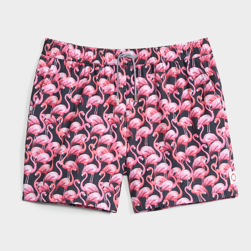 The PINK MIRAGE swim trunks by PUBLIC BEACH SWIM feature a pink flamingo print on a dark background, an elastic waistband with drawstring, and quick-dry fabric for comfort both in and out of the water.