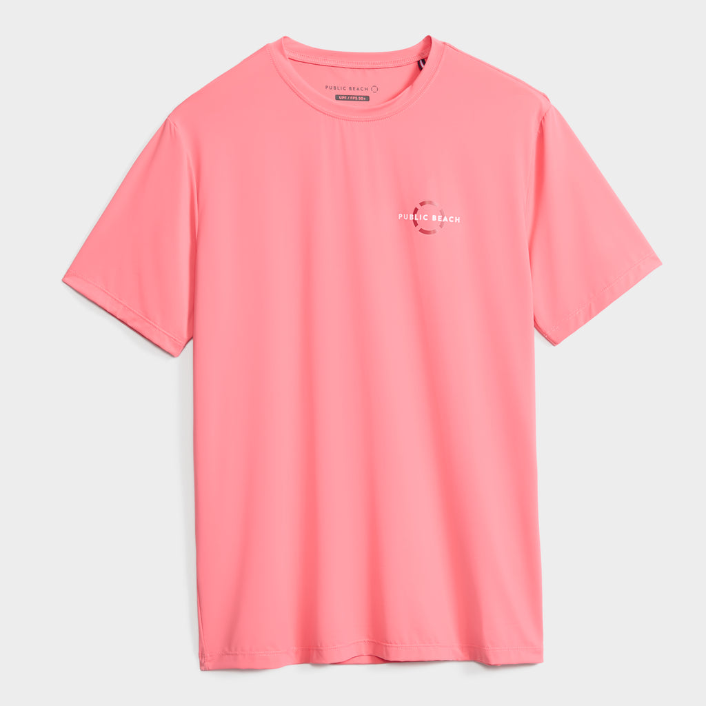 The PUBLIC BEACH SWIM SHORT SLEEVE RASHGUARD is a plain pink shirt with a small white wave logo and "Public Beach" on the left chest. It offers UPF 50 sun protection and is shown on a white background.