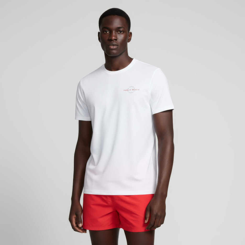 A man with a neutral expression stands against a plain light gray background wearing the PUBLIC BEACH SWIM SHORT SLEEVE RASHGUARD with UPF 50 sun protection and bright red shorts, his arms relaxed at his sides.