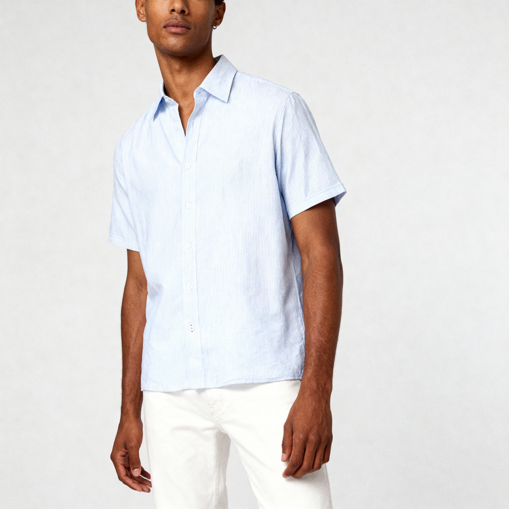 A man stands against a plain light background, facing forward with his left arm relaxed, wearing the PUBLIC BEACH SWIM STRIPE SHIRT—a light blue striped linen short sleeve shirt—paired with white pants.