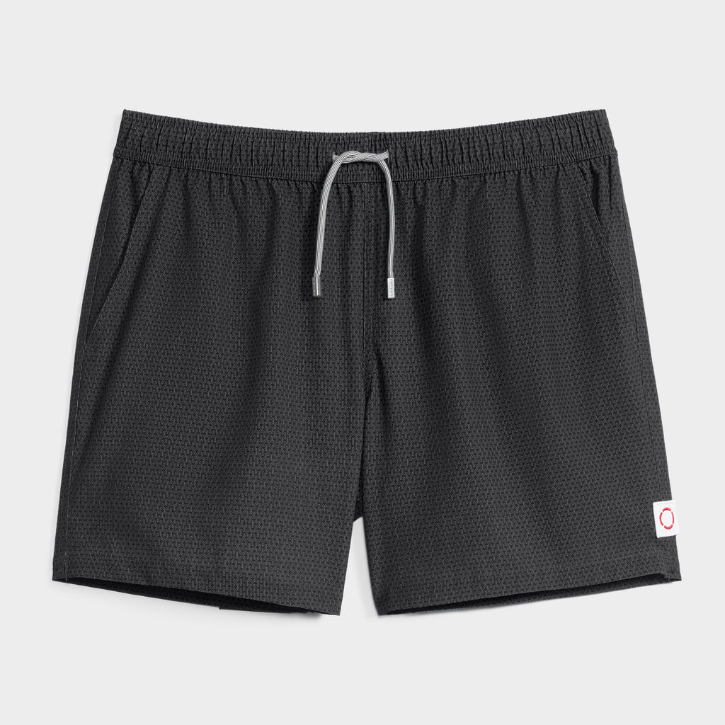 The LINK FLOW by PUBLIC BEACH SWIM is a black mesh men’s swim short with an elastic waistband, gray drawstring, and a small white patch with a red circle on the lower left leg, shown on a plain white background.