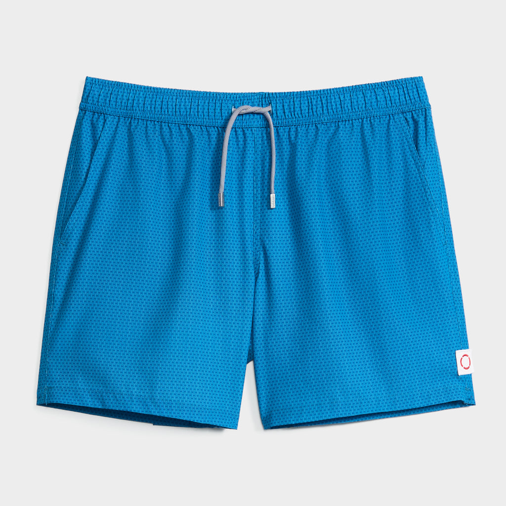 The LINK FLOW swim shorts by PUBLIC BEACH SWIM feature bright blue fabric with a subtle dot pattern, an elastic waistband, gray drawstring, and a white patch with a red circle—quick-dry essentials displayed on a light background.