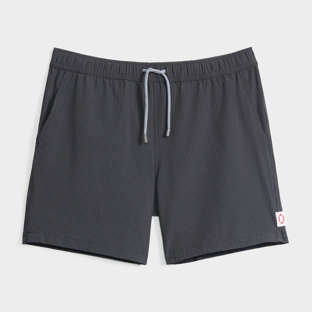 The SAINT TROPEZ men’s swim shorts by PUBLIC BEACH SWIM feature dark gray fabric with subtle vertical pinstripes, an elastic waistband with light gray drawstring, a white tag with red circle, and quick-dry practicality.