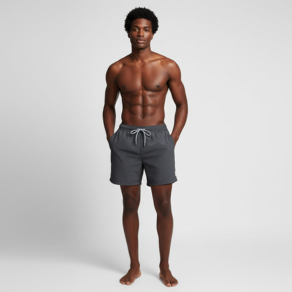 A man stands barefoot against a plain white background, wearing PUBLIC BEACH SWIM's SAINT TROPEZ dark gray quick-dry swim shorts with a drawstring. Hands in his pockets, he looks at the camera, showcasing their comfort and style.