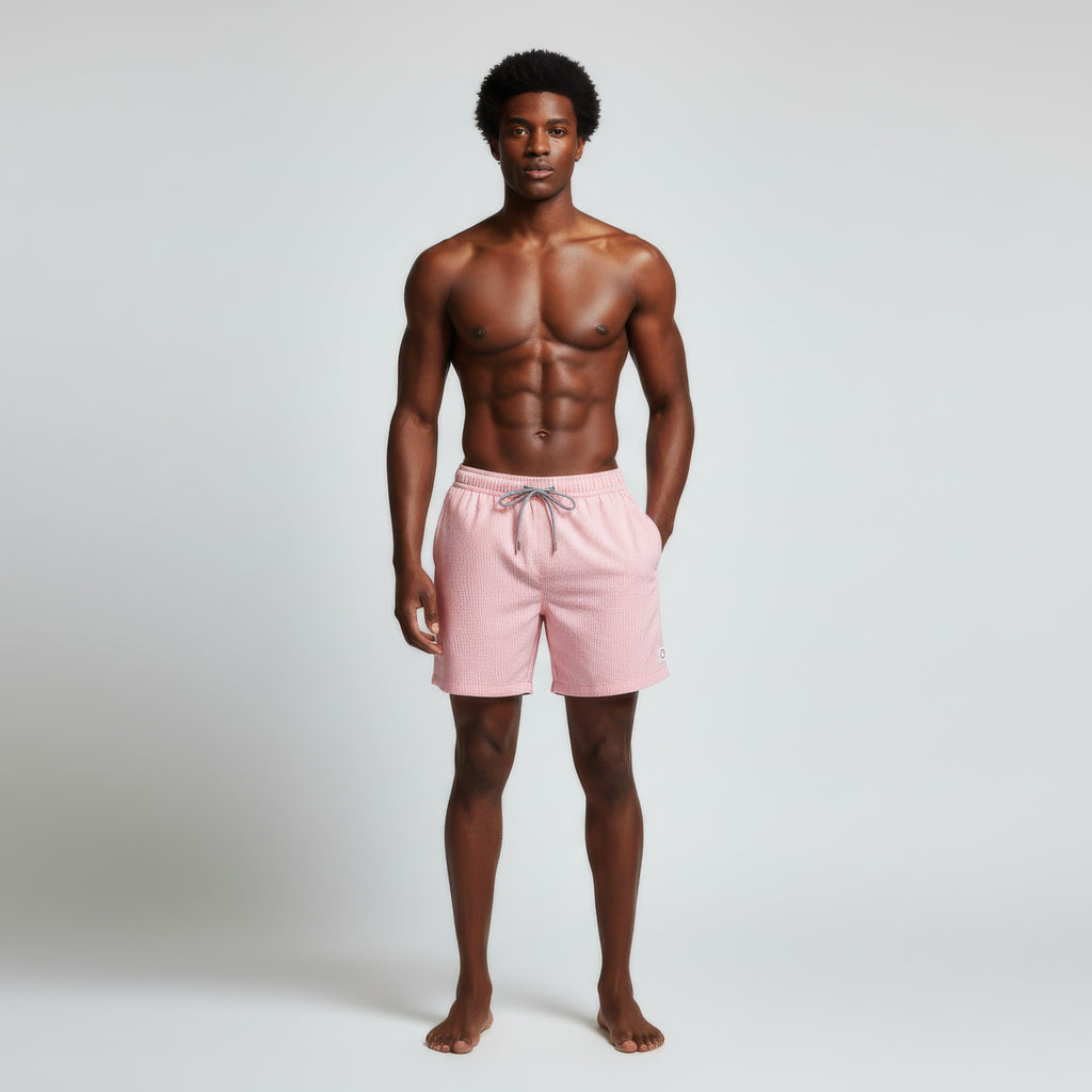 A man stands barefoot against a plain background, wearing PUBLIC BEACH SWIM SAINT TROPEZ pink swim shorts. Shirtless and relaxed, he looks at the camera—showcasing effortless beach style.