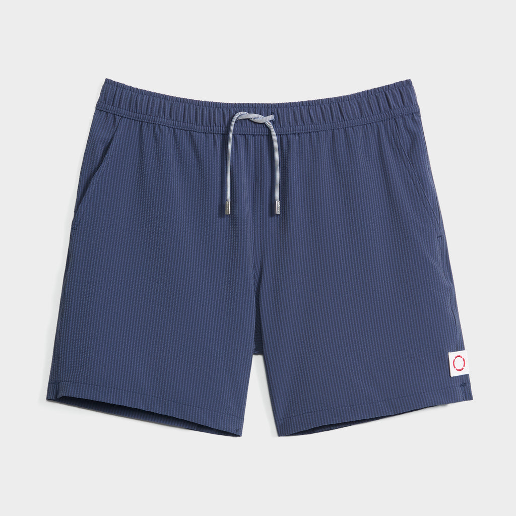 The SAINT TROPEZ men’s swim shorts by PUBLIC BEACH SWIM feature navy blue pinstripes, an elastic waistband with gray drawstring, side pockets, and a small white patch with red circle—quick-dry trunks for comfort and style.