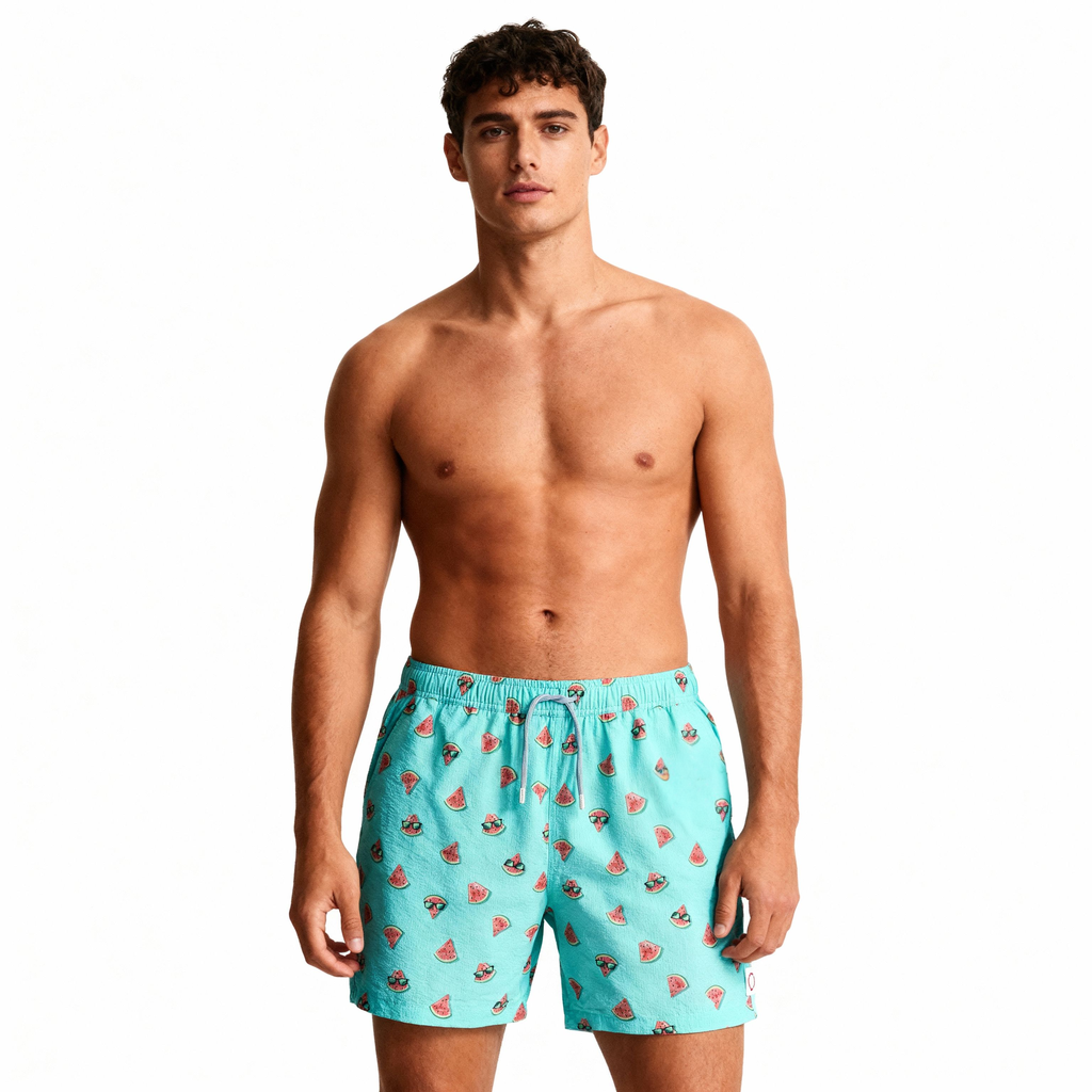 A shirtless man with curly brown hair stands facing forward, modeling PUBLIC BEACH SWIM's SUNNY MELONS light blue swim shorts with a watermelon print against a plain white background.