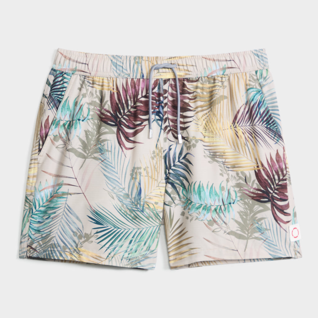 The BOTANIC BURST swim trunks by PUBLIC BEACH SWIM feature a colorful tropical leaf print in green, blue, yellow, and burgundy on light beige, with quick-dry fabric, an elastic waistband, and a drawstring for comfort.