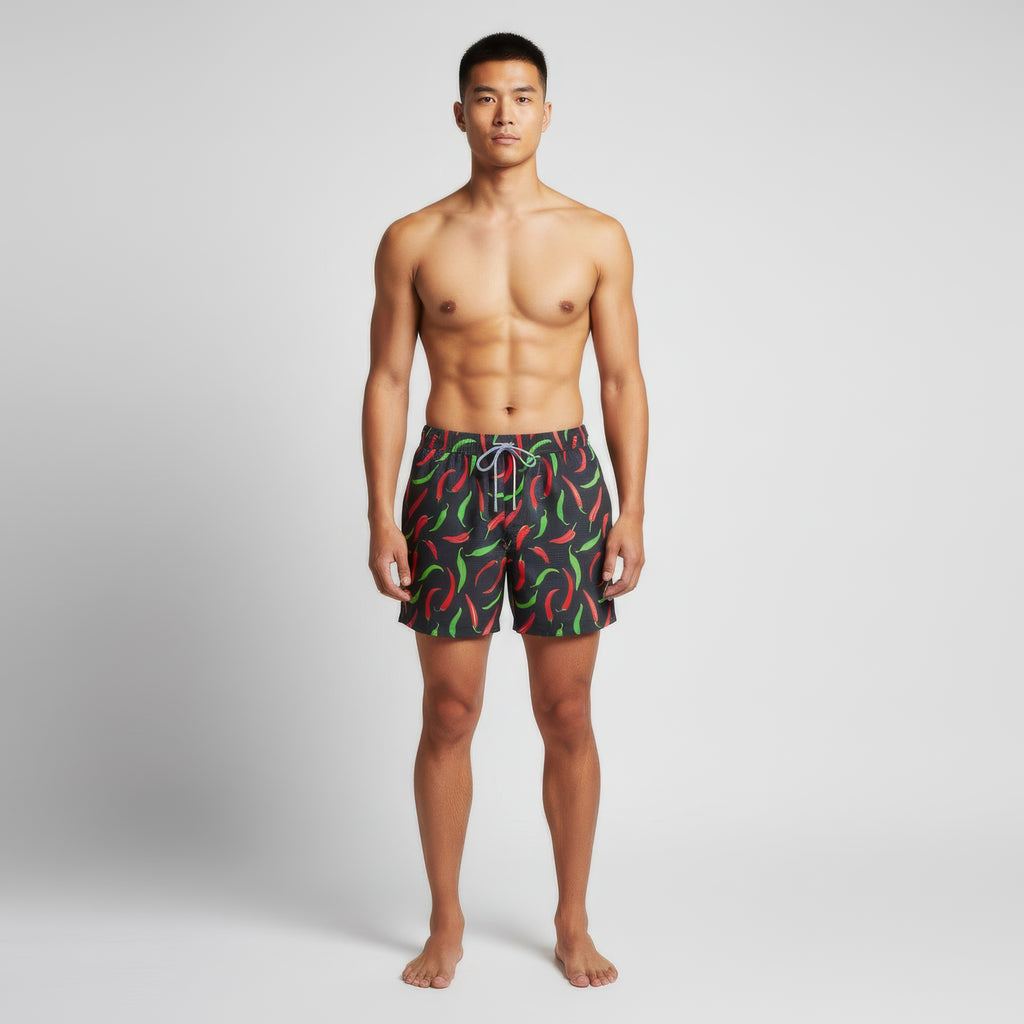 A man stands barefoot against a white background, wearing PUBLIC BEACH SWIM HOT PEPPERS men’s swim shorts in quick-dry fabric with a colorful chili pepper print, facing forward with arms relaxed at his sides.