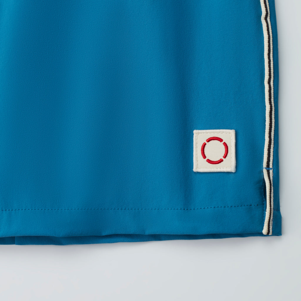 A close-up of the bottom edge of THE VINTAGE swim shorts by PUBLIC BEACH SWIM in blue, featuring white side trim and a small white patch with a red stitched circle—ideal for a classic beachwear look.