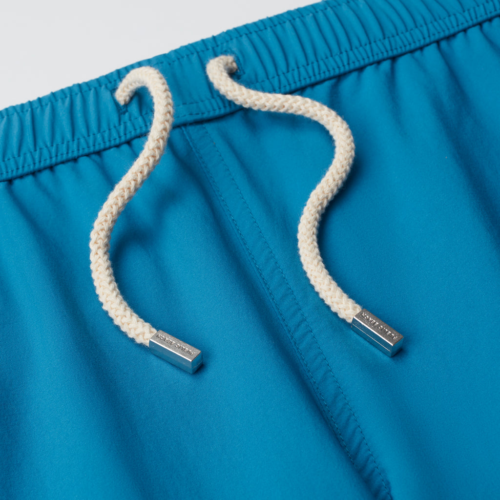 Close-up of THE VINTAGE men's swim shorts by PUBLIC BEACH SWIM in bright blue, featuring white braided cords with silver tips. The elastic waistband and smooth fabric make them perfect beachwear for summer.