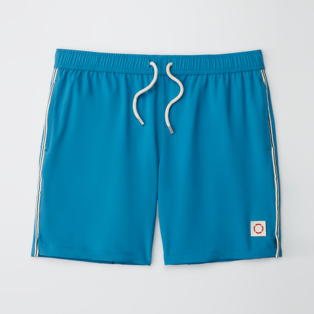 THE VINTAGE men's swim shorts by PUBLIC BEACH SWIM feature bright blue fabric, white drawstrings, white side stripes, and a small white patch with a red circle on the lower left leg, laid flat on a white background.