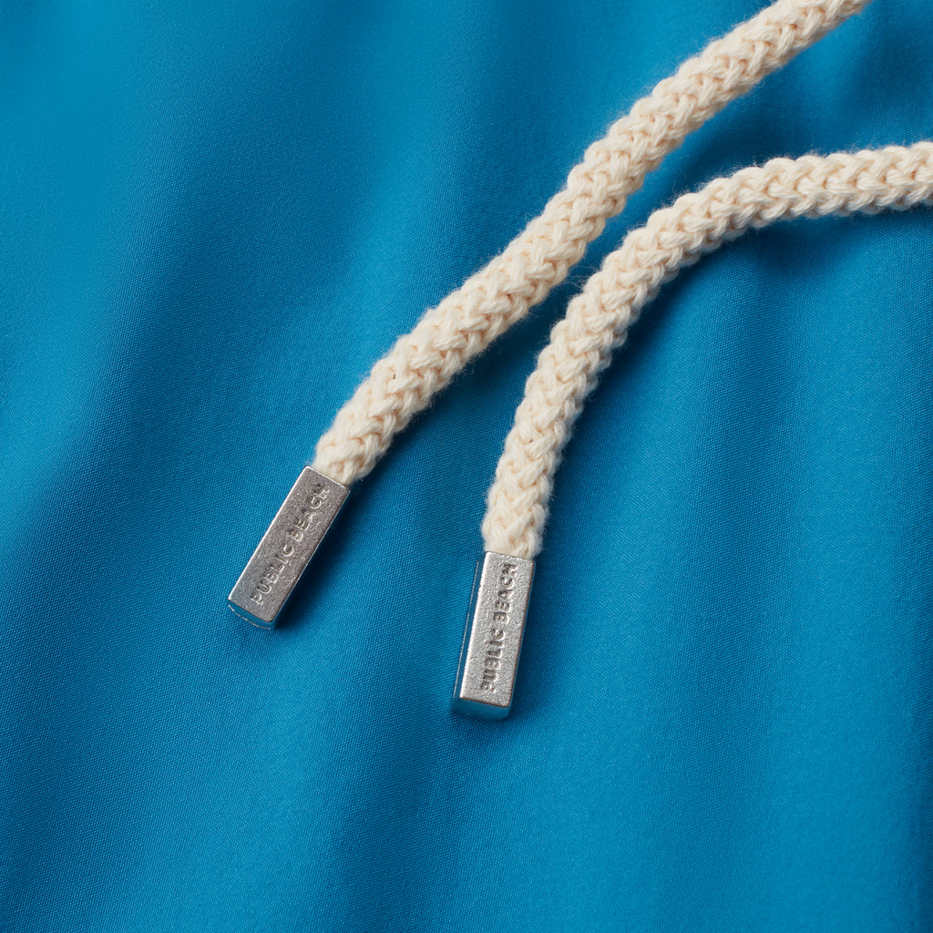Close-up of two white braided drawstrings with silver metal tips, featuring engraved text, on bright blue fabric—ideal for PUBLIC BEACH SWIM's THE VINTAGE men's swim shorts or classic beachwear.
