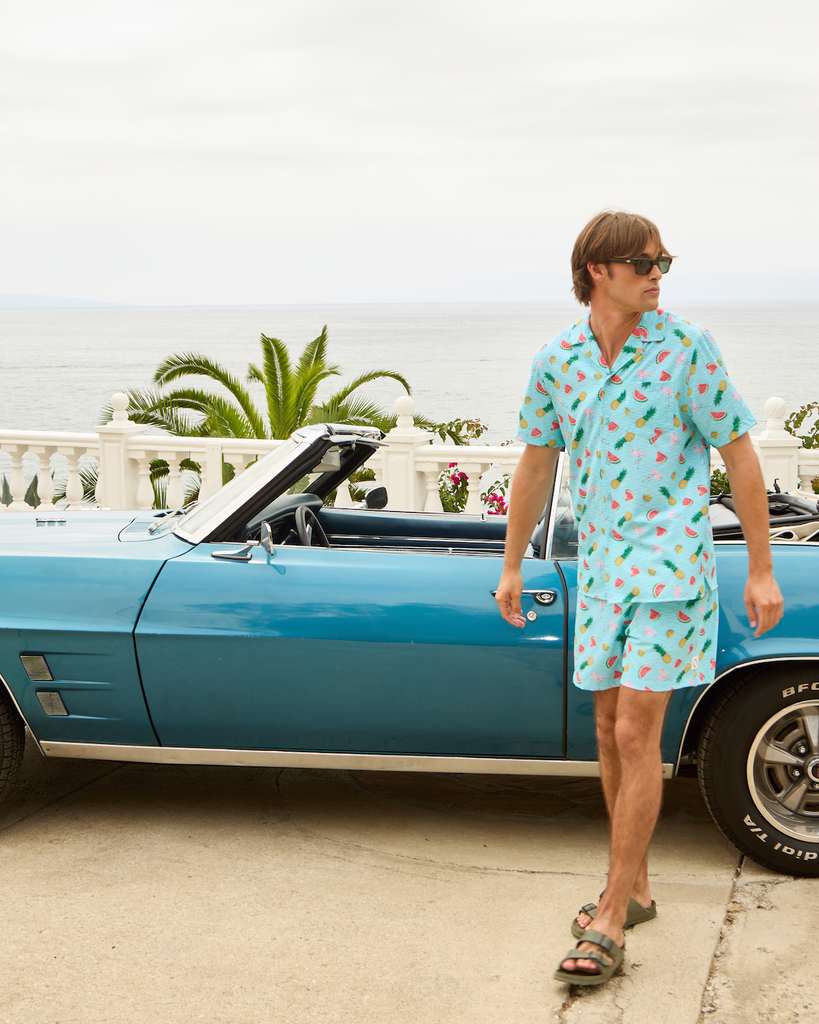 Man in a patterned shirt standing next to a blue classic car with palm trees in the background